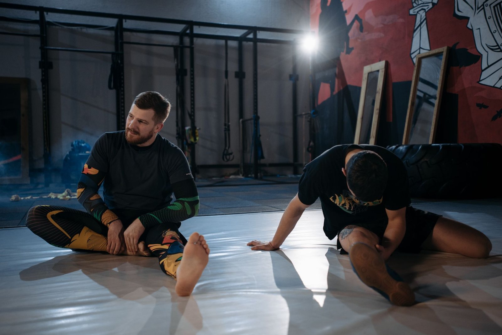 Two men stretching on mats in a gym setting as part of their MMA training routine.