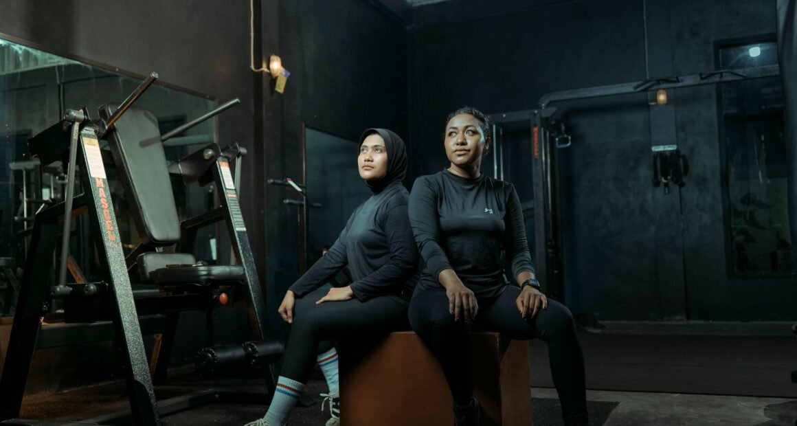 Two women in athletic wear sitting on a box with gym equipment in the background.