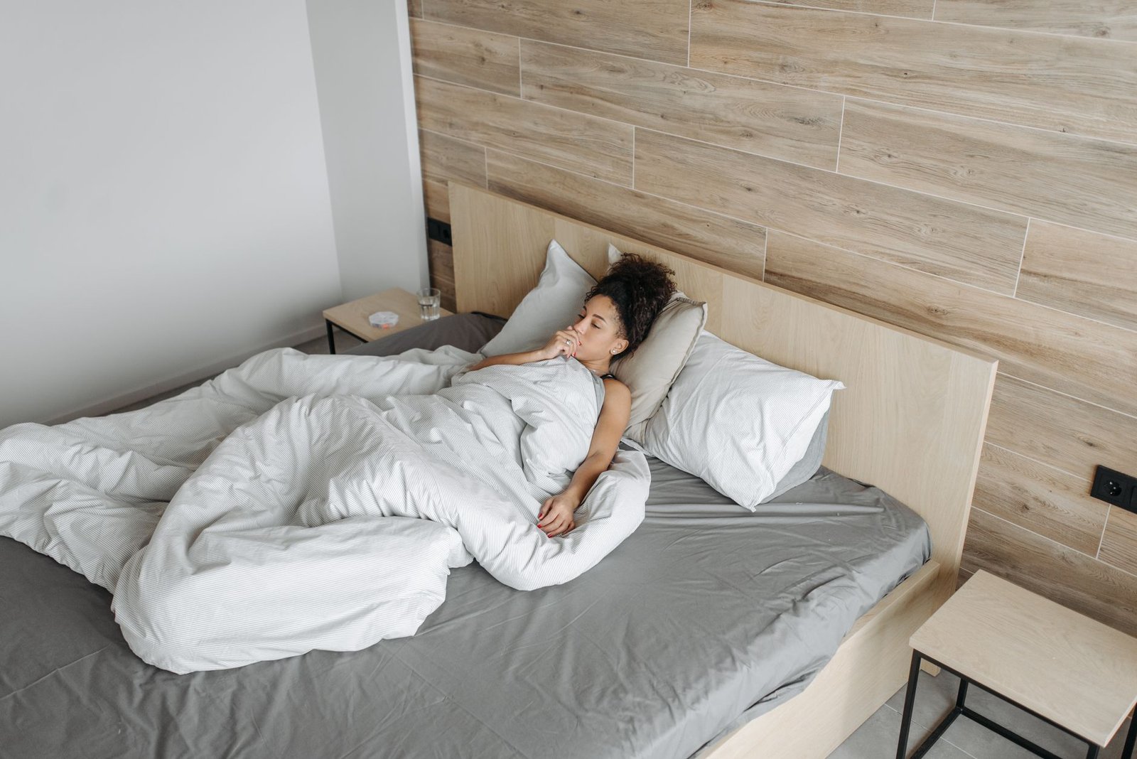 A woman lying in bed under a blanket, appearing unwell, in a modern bedroom setting.